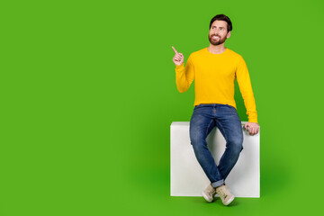 Full length photo of handsome young guy point look empty space sit cube dressed stylish yellow garment isolated on green color background