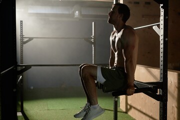 Muscular shirtless man hanging on parallel bars raising legs in static hold to strengthen core...