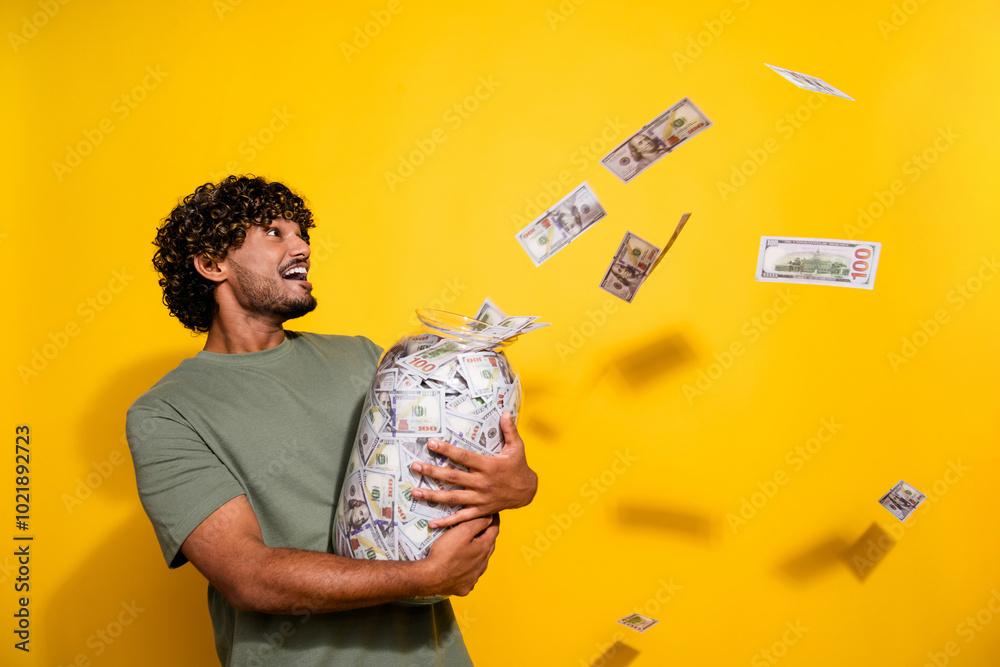 Poster Photo of ecstatic guy with wavy hair dressed khaki t-shirt hold jar with money look at flying dollars isolated on yellow color background