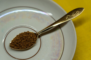 A spoon filled with coffee lies on a saucer on a yellow background. Coffee time
