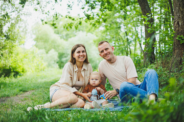 Fototapeta premium A family of a mother and father with a child in their arms, sitting in the park in the summer. The concept of a happy family with a small child, care and love. Happy family with a child.