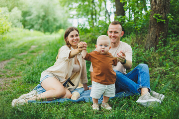 Fototapeta premium A family of a mother and father with a child in their arms, sitting in the park in the summer. The concept of a happy family with a small child, care and love. Happy family with a child.