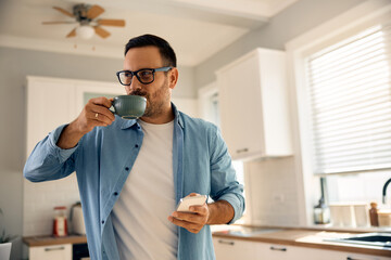 Smiling man drinking coffee while using cell phone in kitchen.