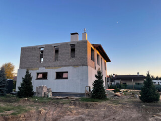 A house is being built in the countryside, featuring a partially completed structure on a plot with surrounding earth and construction materials in the evening light