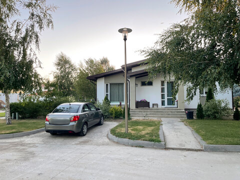 A quaint countryside house is set against a serene backdrop, featuring a parked car in the driveway. The evening light casts a warm glow on the well-maintained yard