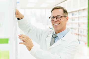 Smiling helpful caucasian mature middle-aged pharmacist druggist looking at camera while searching looking for pills, painkillers, vitamins, antibiotics at pharmacy drugstore