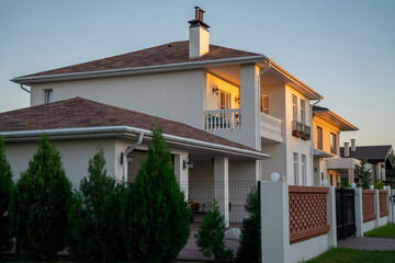 A beautifully designed cottage village house stands proudly with a well-manicured lawn and vibrant flowers. The late afternoon sun casts a warm glow, enhancing its inviting appearance