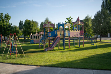 A vibrant playground features slides and climbing structures, surrounded by lush trees and grassy areas, where children enjoy playtime under the sun