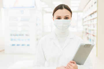 Caucasian female druggist chemist pharmacist in medical mask against pandemic coronavirus holding clipboard looking at camera in chemist`s shop