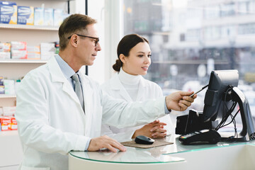 Young caucasian female pharmacist pointing showing male colleague medication prices, prescriptions...