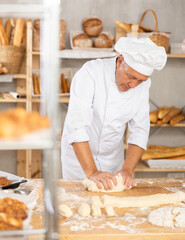 Elderly male baker in uniform kneading raw dough on table
