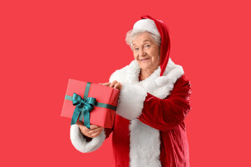 Senior woman dressed as Santa Claus with Christmas gift on red background