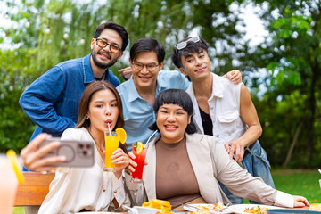 Group of Happy Asian millennial people friends enjoy and fun celebration reunion meeting using mobile phone taking picture together at dinner party in the garden at restaurant on holiday vacation.