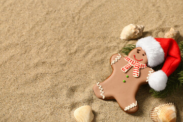 Tasty gingerbread cookie with Santa hat, fir twigs and sea shells on sand background