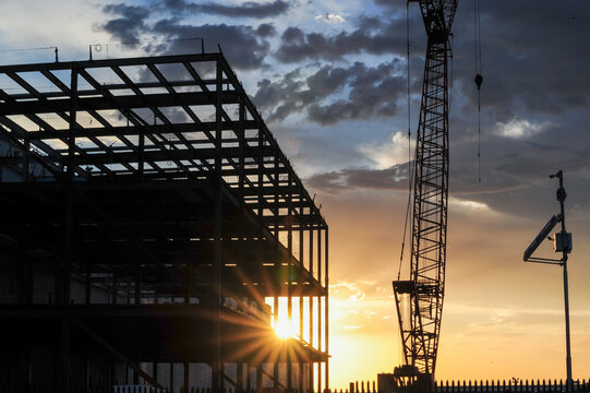 Sunset light outlines the steel beams and tower crane of a large commercial building under construction against a dark blue, cloudy sky.