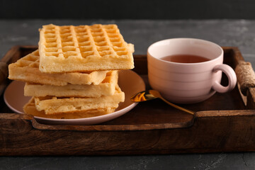 Wooden tray with tasty Belgian waffles and cup of tea on black background