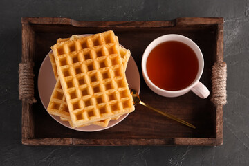 Wooden tray with delicious Belgian waffles and cup of tea on black background