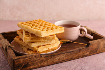 Wooden tray with delicious Belgian waffles and cup of tea on pink background