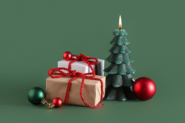 Gift boxes with candle in shape of Christmas tree and balls on green background
