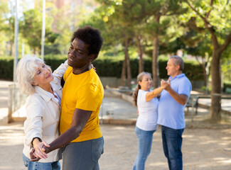 Fototapeta premium Exited older and middle-aged diverse people wearing casual clothes while enjoying latin dances breaking into pairs on sandy area between trees