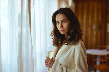 Young woman in robe holding glass of sparkling wine in front of lit window in cabin