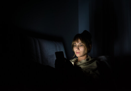 young woman staring at cell phone with face illuminated by screen late at night on sofa