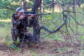 Hunter in camouflage clothing is in kneeling position behind the tree and aiming his rifle at game...