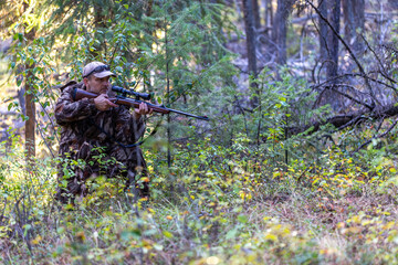 Hunter in camouflage clothing in a half-kneeling position, holding a rifle while slowly approaching...