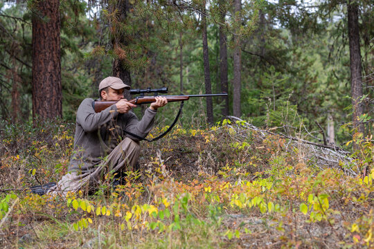Hunter in kneeling position aiming his rifle at game while looking through the scope.  Ready to shoot.  Side view.