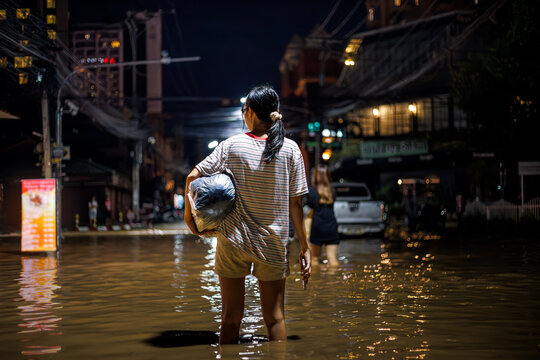 flood victims woman stands in the flooded streets of Chiang Mai city at night 2024, holding a bag with personal belongings. Another person wades ahead through the water