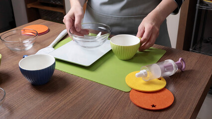 Process of preparing scales and utensils to prepare the dough. Woman removes weight of containers for weighing food. Scales, utensils on wooden table. Baking macarons at home. Hands close-up.