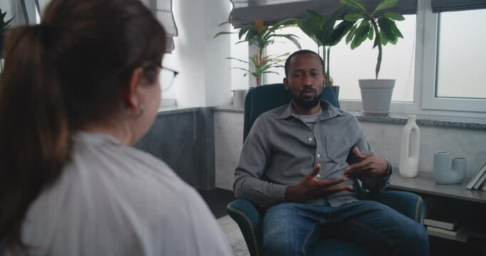 African American adult man sits in chair, talks to female therapist, discuses problems during therapy. Back view of psychologist listening patient, conducting counseling session. Mental health concept