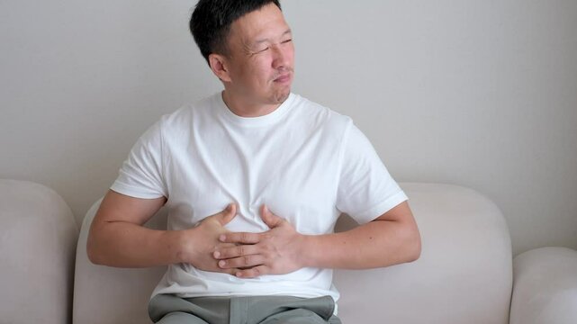 Young man sitting on sofa holding his stomach with hands, concept of indigestion.