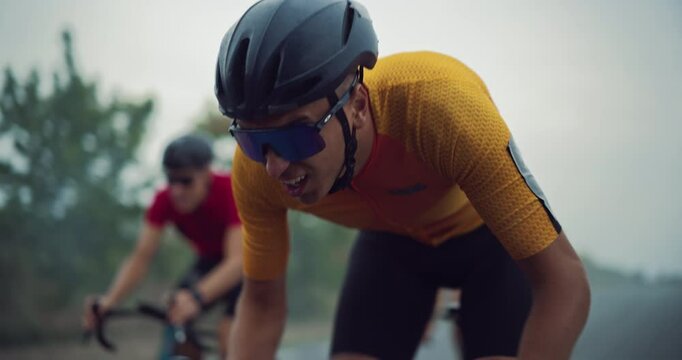 Close Up Portrait of a Cyclist in an Orange and Yellow Jersey Racing on a Misty Road Against Competitors. Intense Dynamic Focus and Pedaling Highlight the Thrill of Cycling Outdoors