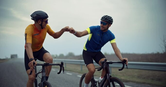 Professional Athletes Riding Together on Bikes, Glancing at Each Other and Offering a Quick Fist Bump, Celebrating Their Shared Effort as They Continue Moving Along the Road. Slow Motion