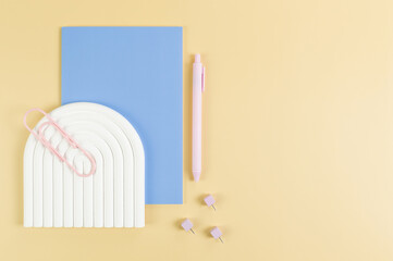 Top view of blue notebook, pink pen, paperclip, pins, white arch tray on yellow background. School, office wallpaper. Flat lay, copy space.