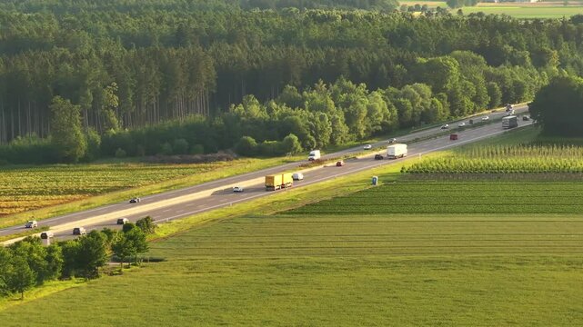 Cars and trucks drive on Germany's A8 highway, surrounded by green fields and forests at sunset