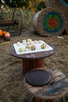 Round wooden table set outdoors on a straw-covered ground with tic-tac-toe board made of white material. Small pumpkins are used as game pieces