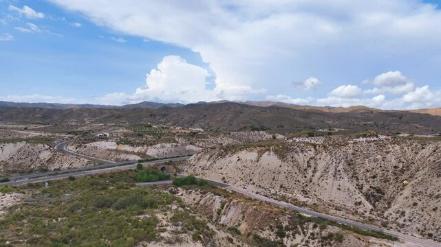 Aerial cruises over rugged and arid inland Spanish landscapes towards a two lane highway bisecting the environment. Tall sculptural clouds dominate the horizon over the vast hilly horizon Sorbas
