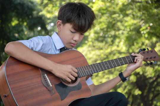 Asian schoolboy in school uniform holding ukulele, sitting in school park and playing it, soft focus.