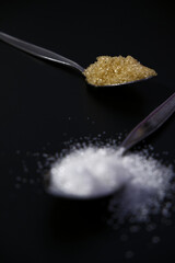 Brown and white sugar in spoons on a black background. Selective focus on brown sugar and blurred in front. Sweet, unhealthy, spilled, and eating concepts. Vertical sugar on spoons with a dark theme.