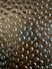 a surface with a bumpy, pebbled texture, possibly resembling droplets or bubbles on glass. The texture creates a reflective and abstract pattern, distorting light and objects behind