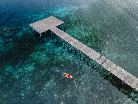 Woman swimming in blue ocean near wooden pier on her vacations. Aerial view - Powered by Adobe