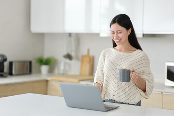 Happy asian woman using laptop drinking coffee in the kitchen