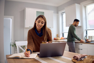 Smiling woman taking notes while using laptop in kitchen.