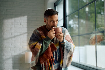 Man wrapped in  blanket drinking tea by window.