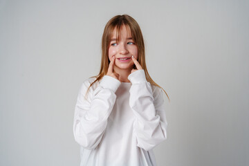 A young girl wearing a white long-sleeve shirt smiles and playfully poses with her fingers on her cheeks in a studio