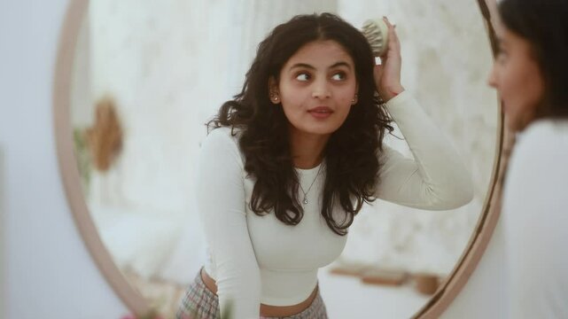 Portrait of pretty dark haired indian woman looking at mirror reflection and doing self hair scalp massage with massager at home Concept of hair loss growth stimulation trichology self care	