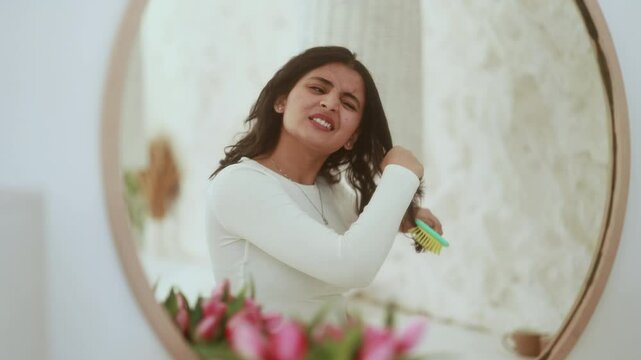 Nervous indian young woman having difficulty brushing her tangled hair dryness split ends problem by wooden brash while looking at mirror reflection at home	