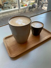 Two cups of coffee, large latte and small cortado on top of a wooden tray at a cafe table in front of a window. Morning coffee window people watching. 